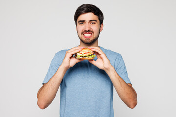 Young dissatisfied disappointed sad man wear blue t-shirt casual clothes hold eat burger look...