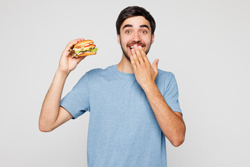 Young shocked surprised man wear blue t-shirt casual clothes hold eat bite burger cover mouth with...