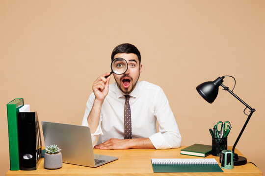 Young shocked successful employee business man wear white shirt tie sit work at office desk with pc laptop look through magnifier loupe isolated on plain beige background. Achievement career concept. - Powered by Adobe