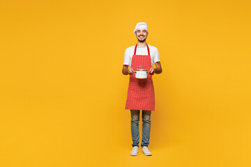 Full body young happy housewife housekeeper chef cook baker man wear red apron toque hat hold in hand saucepan stand look camera isolated on plain yellow orange background studio Cooking food concept