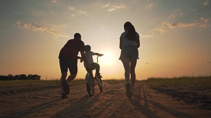 Boy rides bike across dirt road while mother follows close behind. Sunset shines in distance. Outdoor ride gives child freedom. Mother offers training help, child enjoys every ride moment with support