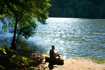 Bearded man sitting on log by lake drinking coffee on sunny day. Outdoor relaxation and travel lifestyle concept