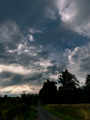Fototapeta premium Starker Abendhimmel, dunkle Wolken, Wald und Weg an Feldern