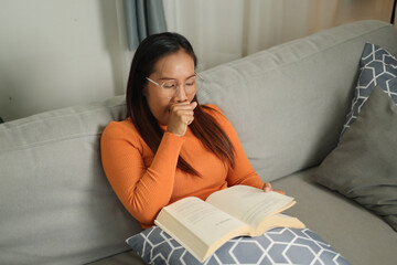 High angle view shot, Asian woman wearing glasses looks relaxed and enjoys reading a book on a comfortable sofa in a living room.