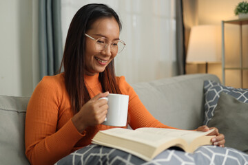 Portrait of Asian woman wearing glasses looks relaxed and enjoys reading a book on a comfortable...