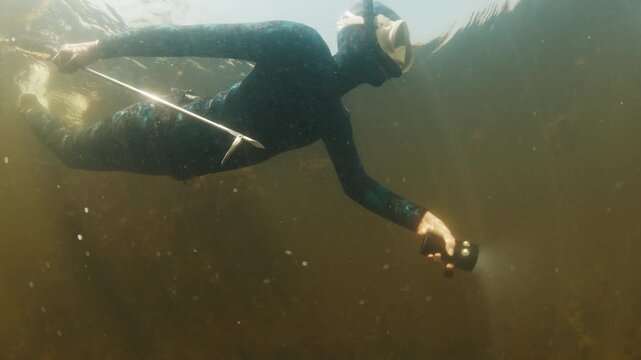 River spearfishing. Woman with speargun and torch slowly swims underwater in the murky river and explores area full of underwater plants