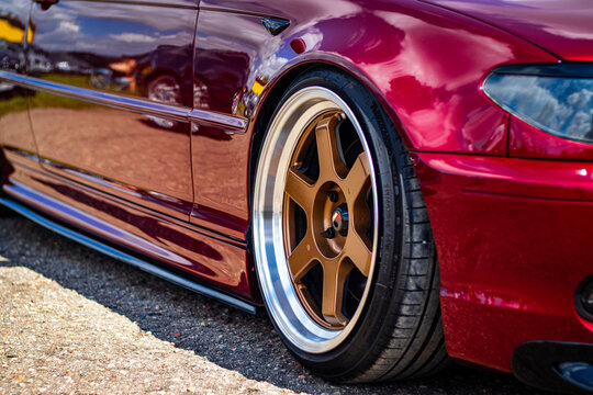 Close-up of a modified red car featuring bronze aftermarket wheels and low-profile tires, reflecting surroundings in its polished body.