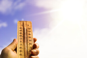 One hand holds a wooden thermometer against a background of sky and bright sunlight indicating a...