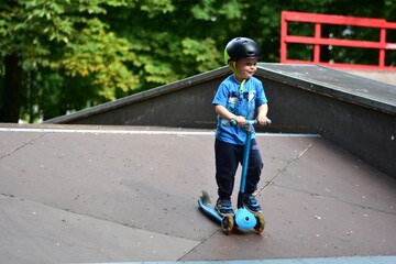 Young boy in helmet rides blue scooter on skate ramp, looking focused as he moves downward. Lush...