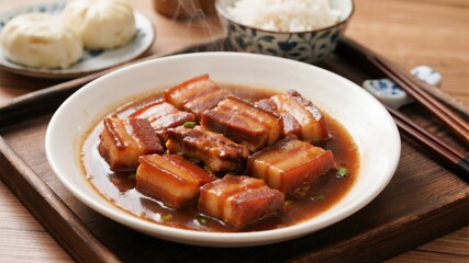 Steaming hot pork belly, distinct chunks of pork belly, served on a white deep plate with rice or steamed buns on the side, with chopsticks and a blue and white porcelain bowl in the background.
