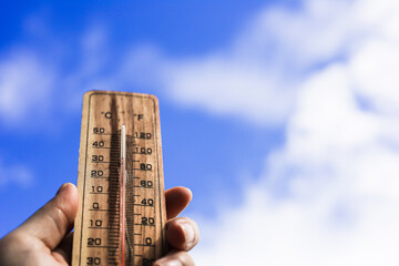 One hand holds a wooden thermometer against the sky background indicating a high temperature.