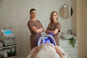 Confident estheticians standing armscrossed beside patient under blue led facial therapy in spa