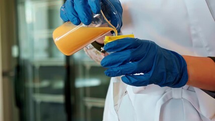 A close-up shot shows a scientist in a lab coat and blue gloves carefully pouring an orange liquid from a glass beaker for a medical or chemical research experiment. - Powered by Adobe