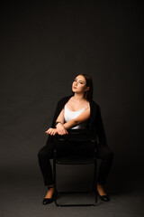 Woman in black pantsuit and white top on black background in studio sitting on black folding chair