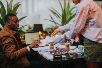 Business colleagues collaborating over documents with charts, analyzing strategies and sharing ideas in a creative workplace setting surrounded by plants and lattes.