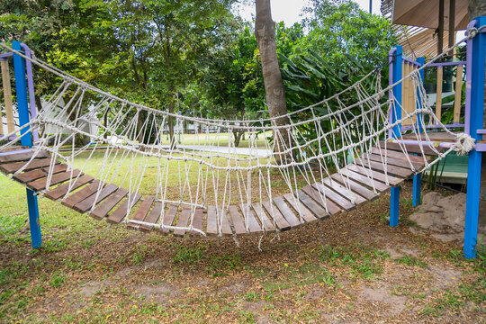 Fototapeta Suspension rope bridge with wooden planks in a playground surrounded by lush green trees and grass on a sunny day