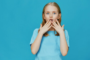Surprised young girl in a light blue shirt, expressing shock with hands on her face against a...