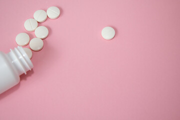 White round tablets are spilling from a white bottle onto a soft pink background, creating a...