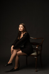 A woman in a short black dress on a dark background in a studio. She has long blonde hair. She is...