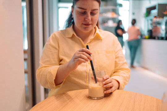 A woman in a yellow shirt stirs an iced coffee drink with a black straw while sitting at a wooden table in a bright, contemporary cafe. The atmosphere is relaxed and minimalistic, with soft natural