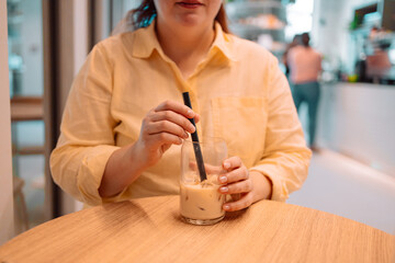 A woman in a yellow shirt stirs an iced coffee drink with a black straw while sitting at a wooden table in a bright, contemporary cafe. The atmosphere is relaxed and minimalistic, with soft natural