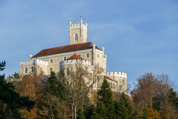 A close-up view captures the upper portion of Trakošćan Castle, highlighting its main tower and battlements. The historic structure rises above the surrounding treetops against a clear blue sky.