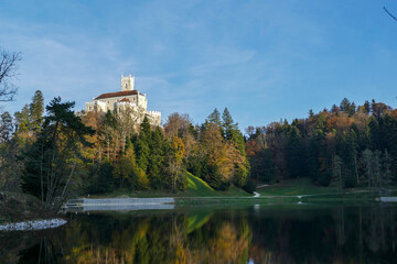 Trakošćan Castle is seen from across a calm lake, nestled atop a hill covered in colorful autumn forests. The picturesque scene is reflected in the still water below under a vast blue sky.