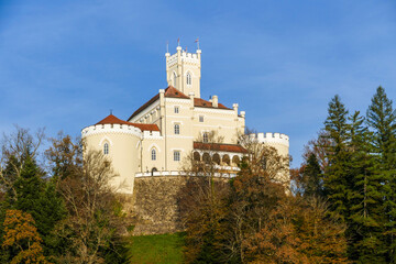 Trakošćan Castle sits atop a sun-drenched hill with vibrant green grass and autumn-colored trees. Below, the calm lake water provides a perfect, mirror-like reflection of the forest and hillside.