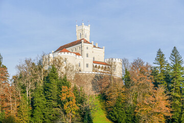 A majestic, cream-colored castle with red roofs and a central tower sits atop a wooded hill. The surrounding trees show autumn colors under a clear, bright blue sky.