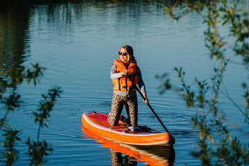 A girl in a life jacket and sunglasses is sitting on a paddleboard floating on the river.