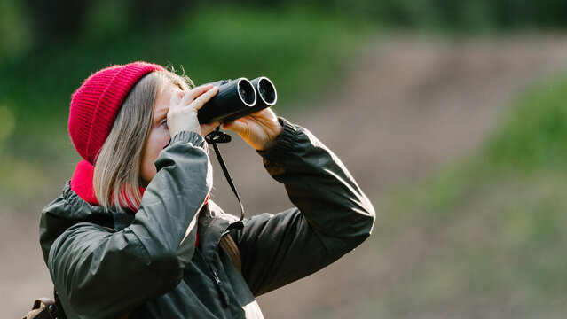 A middle-aged girl with binoculars in a red hat and a green jacket stands against the background of a forest road and looks into the distance.