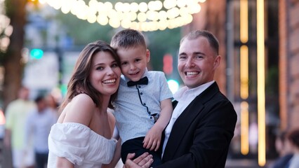 Father, mother and son laugh and hug on the street during city festival at sunset. Happy family in the evening.