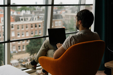 young man working on laptop in a chair with attic window and view on Amsterdam	