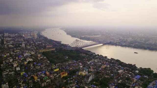 A cinematic daylight drone aerial shot of the historic or iconic Howrah Bridge over holy Hooghly or Ganges river, surrounded by Kolkata or Calcutta city with a vibrant cloudy sky in the background.