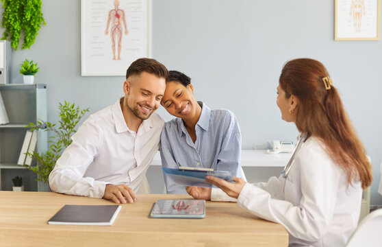 Happy young couple sitting at desk and talking with family doctor therapist looking to report file with appointment during medical exam in clinic planning pregnancy. Health care concept.