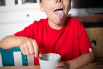 Child enjoys eating yogurt in a cup while sticking out tongue playfully in a bright home with...