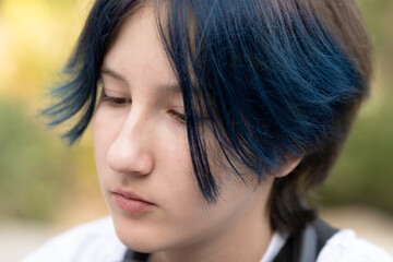 Close-up portrait of a thoughtful teenager with blue hair looking down outdoors in natural light,...