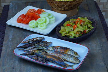 A simple, traditional Indonesian meal featuring fried fish, stir-fried bitter melon, sliced tomatoes and cucumbers, and steamed rice in a woven bamboo container.