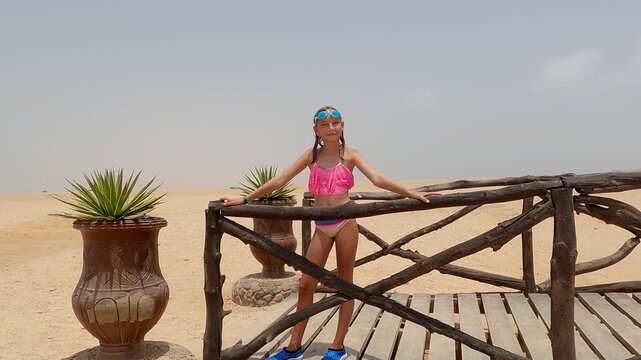 Young girl in pink bikini and blue goggles standing on wooden terrace, overlooking expansive desert landscape with terracotta potted plants adding greenery to arid scene