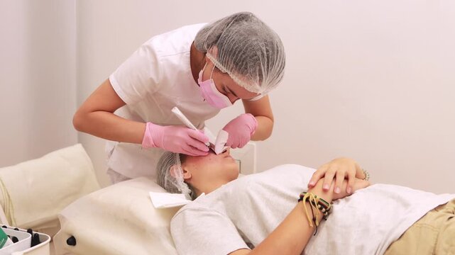 Beautician wearing pink protective gear performing microneedling procedure on female patient's facial skin at medical treatment table. Beauty clinic concept.Teenager facial care concept