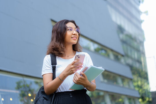 A woman is looking at her phone while holding a note book. She is wearing glasses and a white shirt