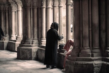 Within the hallowed halls, a figure in a dark cloak lights a candle while another sits quietly, lost in thought against the backdrop of ornate stone pillars at Mosteiro da Batalha