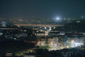At night, buildings are illuminated along a narrow street. A bridge spans a wide river. Faint lights from distant hills twinkle. The sky above is dark, suggesting late hours