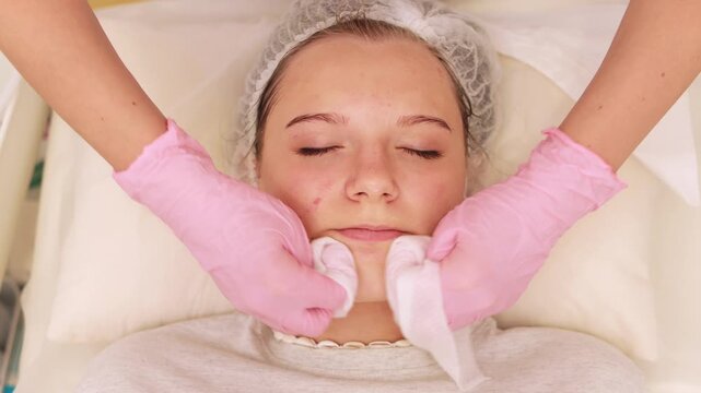 Skincare professional wearing pink gloves cleansing female client's face, removing impurities with cotton pads in pristine medical environment. Beauty clinic concept.Teenager facial care concept