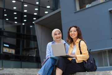Two young women sitting on a ledge with a laptop in front of them.
