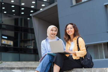 Two young women sitting on a ledge with a laptop in front of them.