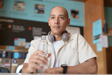 Bald Latin man drinking water in laundromat with washing machines behind
