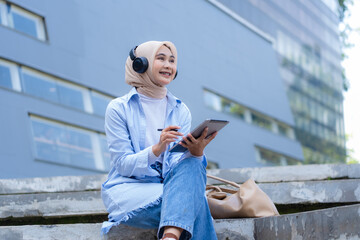 A woman wearing a head scarf is sitting on a ledge and using a tablet. She is smiling and she is...