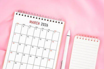 March 2026 month calendar and pen with open diary on pink color fabric background. Monthly calendar concept.