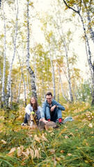 Fototapeta premium A married couple relaxes in nature during a walk with their beloved dog in an autumn forest. The loving woman and man sit in the tall yellow grass, in the fern leaves, with the dog next to them.